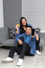Couple of Latin man and woman sitting in their living room using cell phone and credit card to pay, buy with discounts and promotions