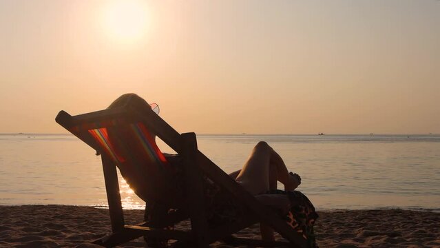 Young woman relaxing in deck chair on tropical sea beach on beautiful sunset. Vacation in Thailand.