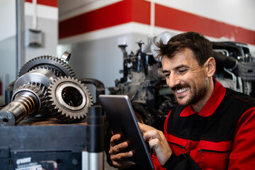 Mechanic typing on tablet computer and standing inside workshop with spare parts.