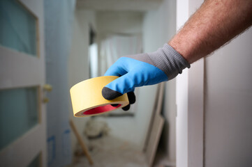 Worker with a masking tape in hands at construction site. Quick and easy painting process. Accessories for install furniture, repair home. Man dressed in blue gloves. Home renovation concept