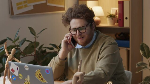 Medium shot of eccentric red-haired male corporate employee sitting at desk with wooden toy figures, in company office, working on laptop, answering phone call for personal conversation during workday
