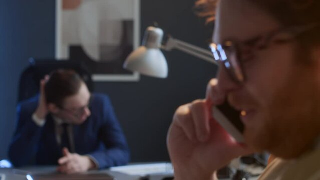 Medium rack focus shot of cheerful man chatting on mobile phone and laughing in office in middle of workday, and angry Caucasian manager sitting at desk, glaring at him and giving nasty looksMedium ra