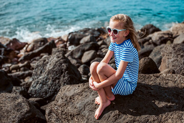 Beautiful girl sitting on rocks by sea. Blonde girl in dress enjoying vacation in Canary Islands. Concept of beach summer vacation with kids.