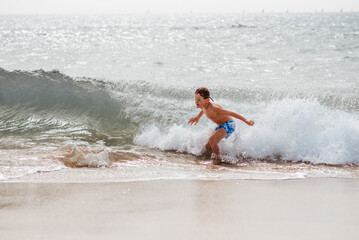 Young boy playing, running and splashing in strong sea waves. Smilling boy in swimsuit looking at waves. Concept of beach summer vacation with kids.