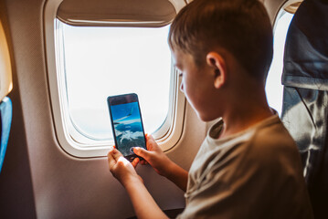 Boy with headphones sitting airplane, taking photos from window. Concept of family beach summer vacation with kids.