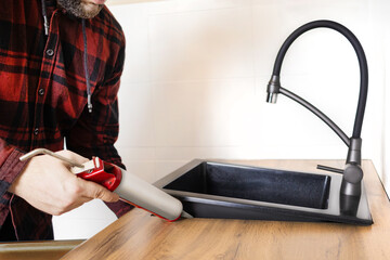 A man installs a black stone sink into a kitchen countertop. Worker seals up the kitchen sink with...