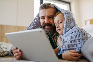 Little boy watching cartoon movie on tablet with father, lying under blanket on floor in kids room. Dad explaining technology to son, digital literacy for kids. - obrazy, fototapety, plakaty