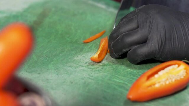 Chef, wearing a blue denim apron and black sterile gloves, skillfully cutting  and slicing fresh Peruvian orange chili peppers into strips on a green plastic cutting board using a sharp chef's knife