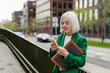 Mature businesswoman scrolling on smartphone, going home from work. Beautiful older woman with gray hair standing on city street.