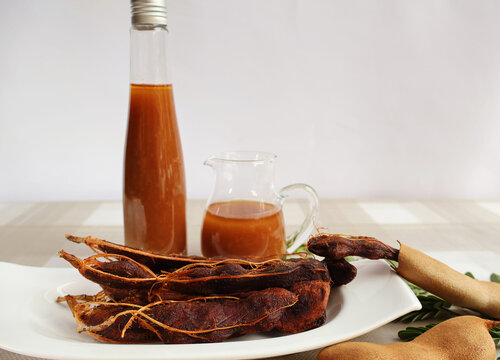 Peeled ripe tamarind fruit placed in a white porcelain plate in front of a bottle and jar of concentrated tamarind juice, white background.

