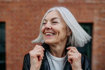 Portrait of stylish mature woman with gray hair on city street. Older woman in leather jacket smiling.