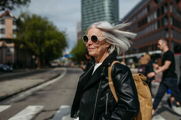 Portrait of stylish mature woman with gray hair on city street. Older woman in sunglasses crossing a road on pedestrian crossing.