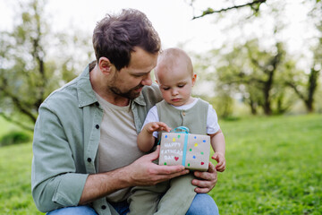 Father holding little baby and gift for Father's day. Family time outdoors in the park during warm spring day.
