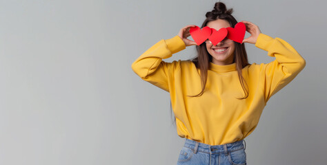 A woman wearing a yellow sweater and jeans is holding up two red hearts in front of her face. Happy woman wearing yellow clear sweatshirt is holding red paper hearts near her eyes in blue jeans - Powered by Adobe