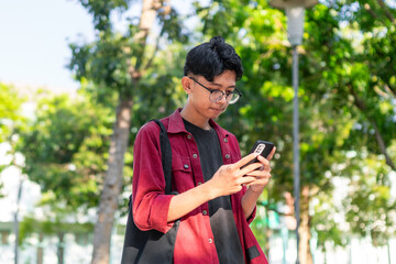 Young Asian college student using smartphone with happy expression. A male smiling while holding his phone and books at the public park. copy space