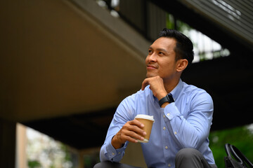 Positive male entrepreneur holding takeaway coffee cup looking away sitting on stairs in the city