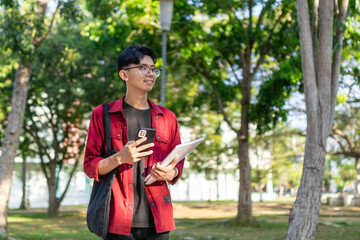 Fototapeta premium Young Asian college student using smartphone with happy expression. A male smiling while holding his phone and books at the public park. copy space