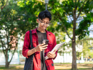 Young Asian college student using smartphone with happy expression. A male smiling while holding his phone and books at the public park. copy space