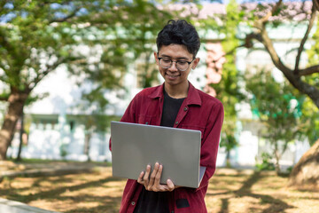 Asian college student sitting and focused to his laptop screen. Asian young male sitting outdoors using laptop computer, typing, working freelance project