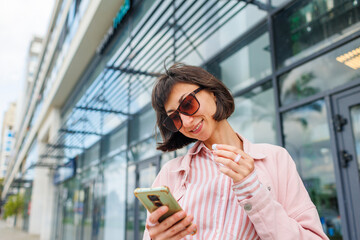 Successful businesswoman in fashionable clothes and sunglasses using wireless headphones outdoors near her office