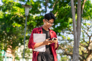 Young Asian college student using smartphone with happy expression. A male smiling while holding his phone and books at the public park. copy space