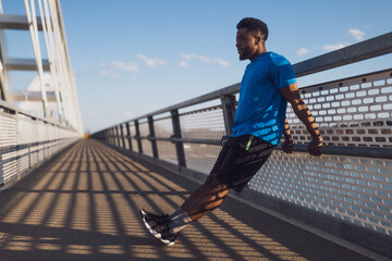 Young african-american man is exercising on the bridge in the city. He is doing reverse push-ups. - 791406240