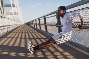 Young african-american man is exercising on the bridge in the city. He is doing reverse push-ups. - 791406243