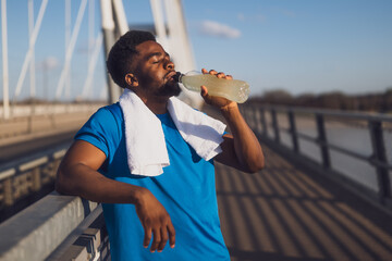 Portrait of young african-american man who is drinking water and relaxing after jogging. - 791406274