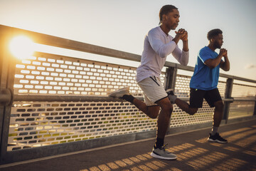 Two african-american friends are exercising on the bridge in the city. They are doing squats. - obrazy, fototapety, plakaty