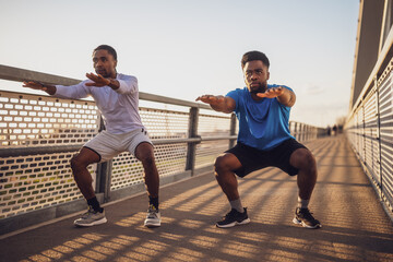 Two african-american friends are exercising on the bridge in the city. They are doing squats.