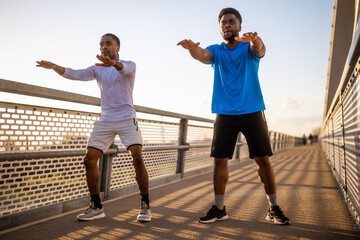 Two african-american friends are exercising on the bridge in the city. They are doing squats. - 791406449