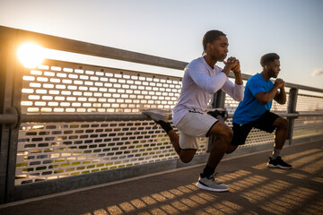 Two african-american friends are exercising on the bridge in the city. They are doing squats. - 791406451