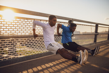 Two african-american friends are exercising on the bridge in the city. They are doing reverse push-ups. - 791406459