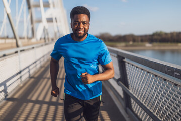 Young african-american man is jogging on the bridge in the city. - obrazy, fototapety, plakaty