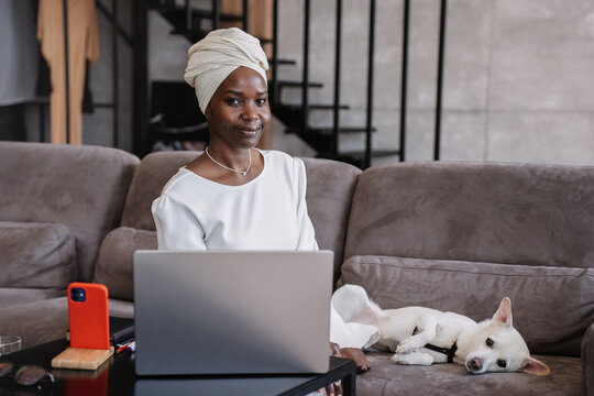 Serene woman with headwrap using laptop on sofa, dog lounging nearby