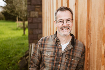 Smiling Senior Man in Glasses Leans Against Wooden Wall Outdoors