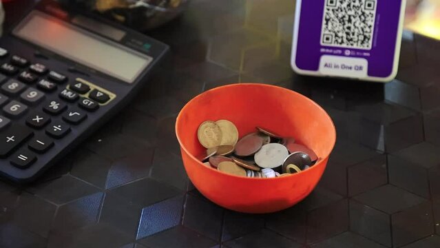 Slow motion shot of Indian coin pouring into bowl filled with coins on a flat surface of grocery shop counter, beside a calculator and QR code payment machine. UPI, wallet payment