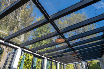 A glass roof of a sunroom creates a light and airy ambiance, allowing natural light to filter through the transparent ceiling, the large windows offer a view of the green trees.