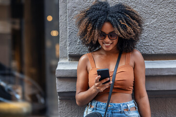 Happy woman using smartphone while carrying shopping bags leaning against the wall in the city