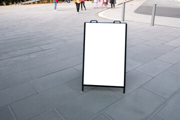 A blank white mockup board on an urban pedestrian walkway, with copy space for design. Background texture of a public advertisement sign or A-frame standing easel displayed in the public.