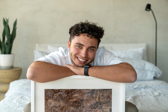 Happy young man sitting with picture frame on bed at home