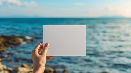 A woman's hand holds a white blank piece of paper against the background of the sea. Mockup, product advertisement, invitation flyer
