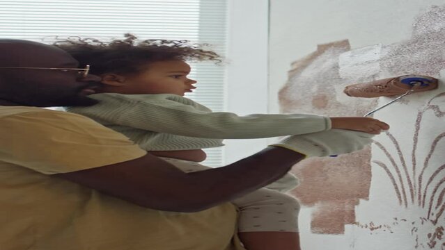Vertical side footage of African American man holding his little Biracial daughter in hands and teaching her how to paint walls with roller during room redecorationVertical side footage of African Ame