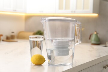 Water filter jug, glass and lemon on white marble table in kitchen