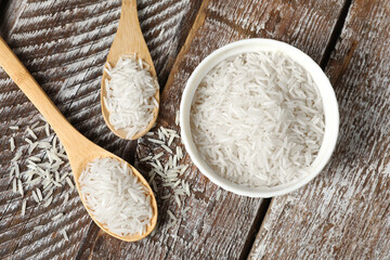 Raw basmati rice in bowl and spoons on wooden table, flat lay