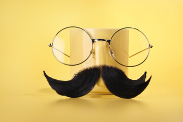 Man's face made of artificial mustache, glasses and cup on yellow background