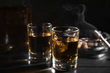 Alcohol addiction. Whiskey in glasses, smoldering cigarette and ashtray on black wooden table, closeup