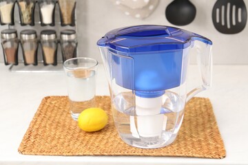Water filter jug, glass and lemon on white countertop in kitchen