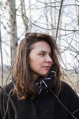 Thoughtful Woman in Black Coat and Geometric Scarf in Front of Bare Tree Branches - 791434247