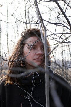 A young woman with long brown hair stands behind a bare tree branch. Serene Atmosphere.